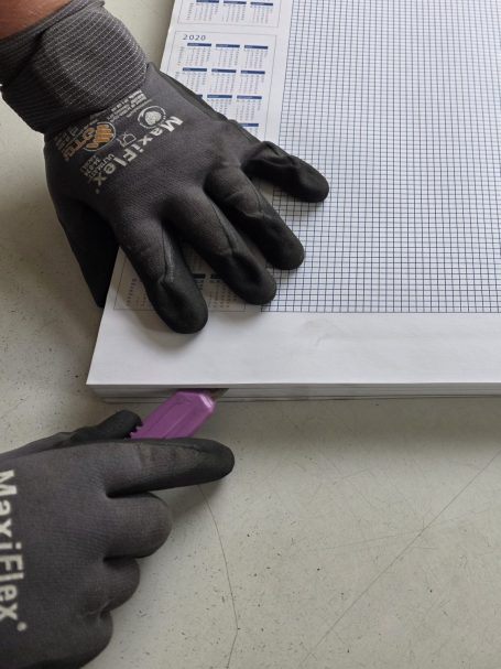 Hände mit Arbeitshandschuhen schneiden eine Kante mit einem Cutter-Messer.